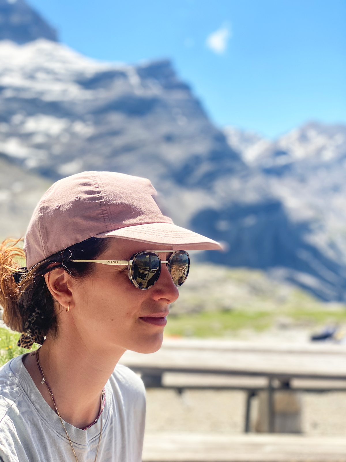 A woman in profile wearing a dusty pink cap and Moiry Tortoise sunglasses, set against a blurred mountain landscape under a blue sky.