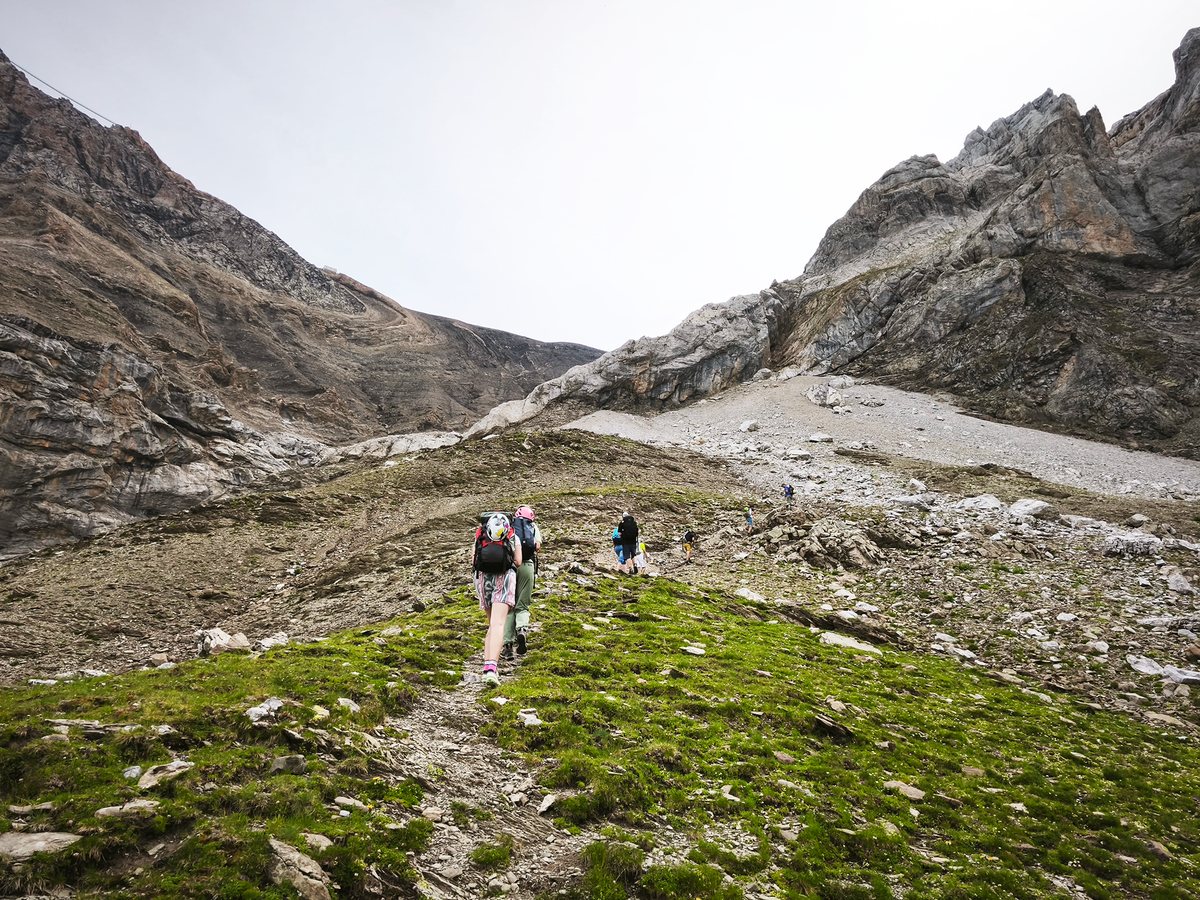 Three hikers with backpacks walk along a dirt trail through green alpine grass, with rocky mountains rising on both sides under an overcast sky.