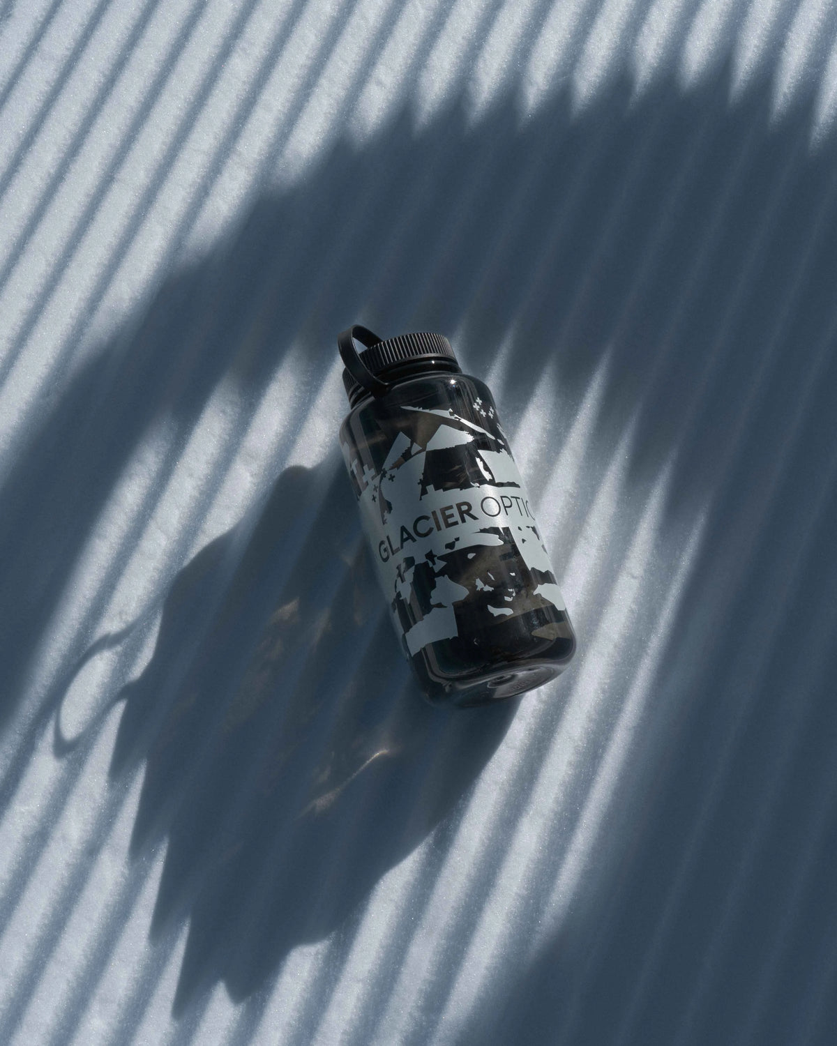 A black and white Glacier Optics water bottle with a mountain logo, resting on a white surface with diagonal shadows.