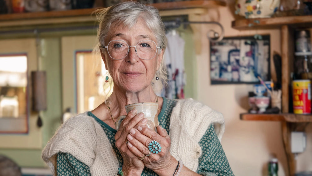 An older woman with grey Laiva optical glasses, holds a ceramic mug with both hands in a cozy, warmly lit kitchen setting.