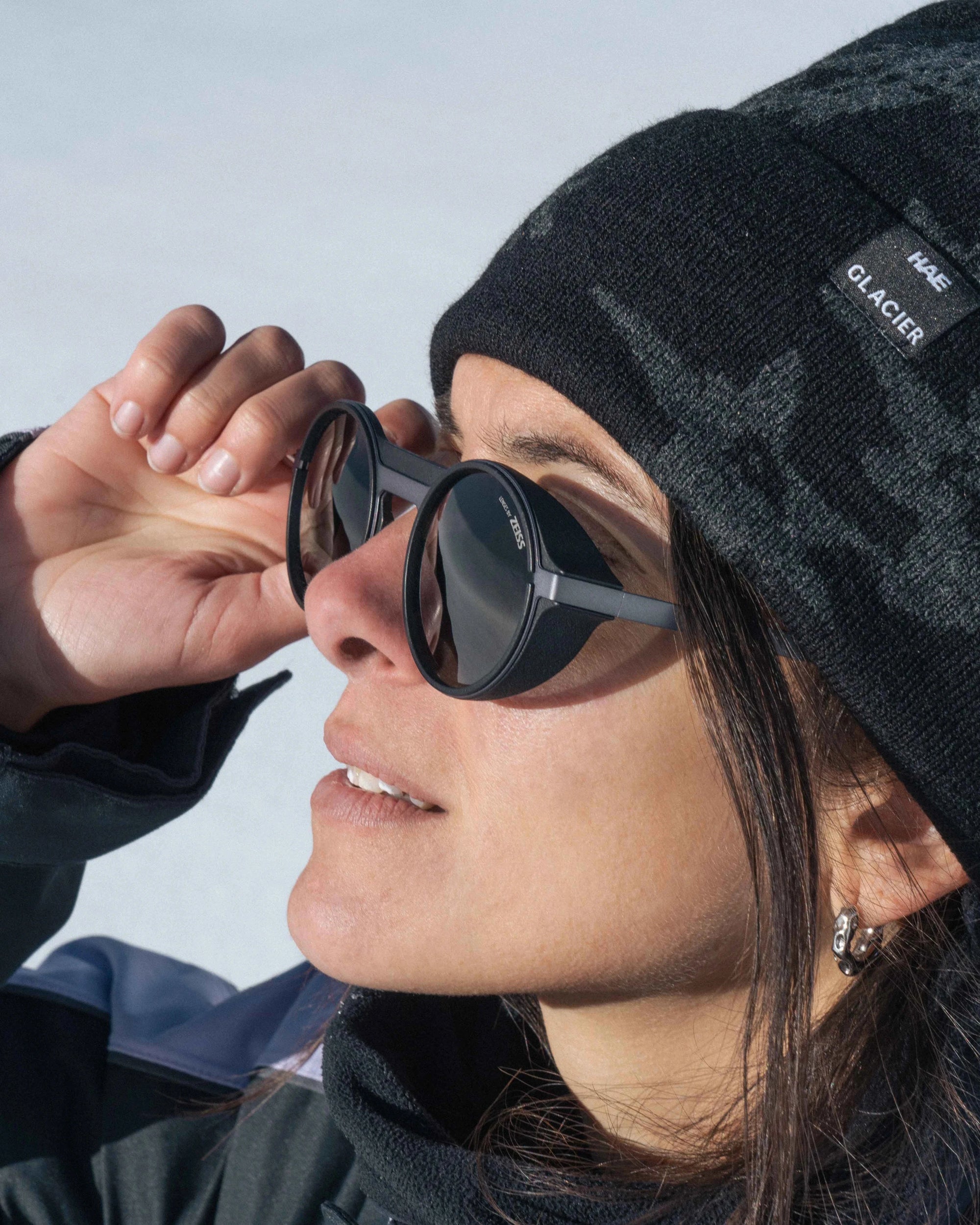 Person wearing a hae x glacier optics black beanie and palü bold grey sunglasses with a clear sky background