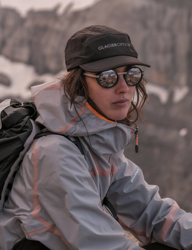 A person outdoors in a mountain setting wearing a Glacier Outdoors cap and palü light silver round sunglasses. 