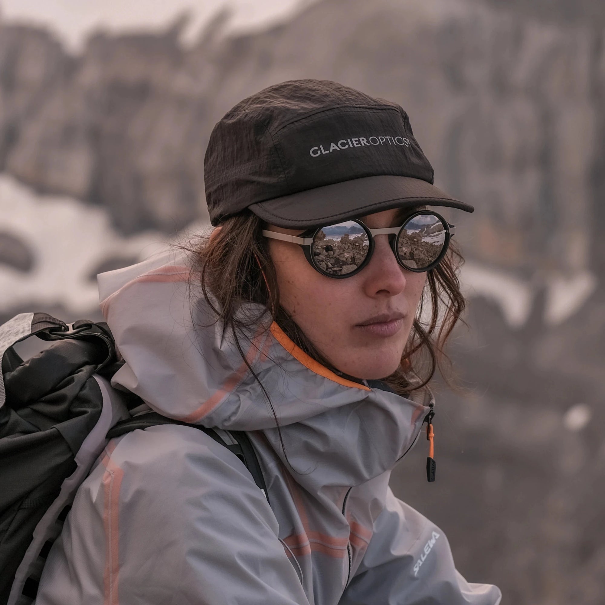A person outdoors in a mountain setting wearing a light gray Glacier Outdoors cap and palü light silver round sunglasses.