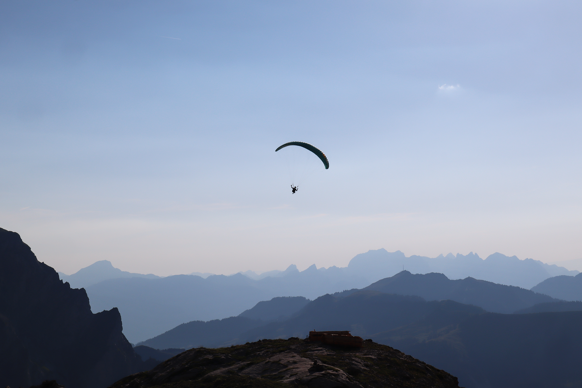 A paraglider hangs in the air above layered mountain ranges under a pale blue sky, with dark rocky peaks in the foreground.