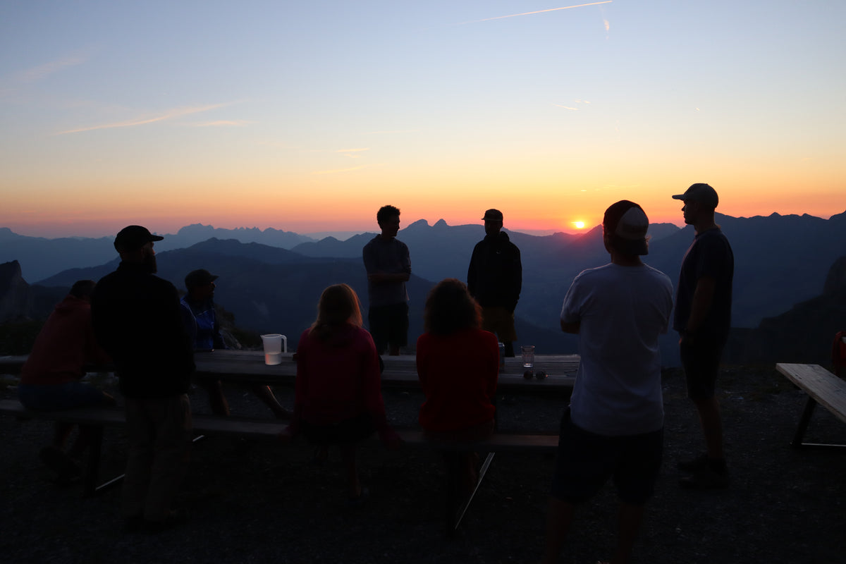 Silhouetted people gathered around picnic tables at a mountain overlook during sunset, with layered mountain ranges and a glowing orange sky in the background.